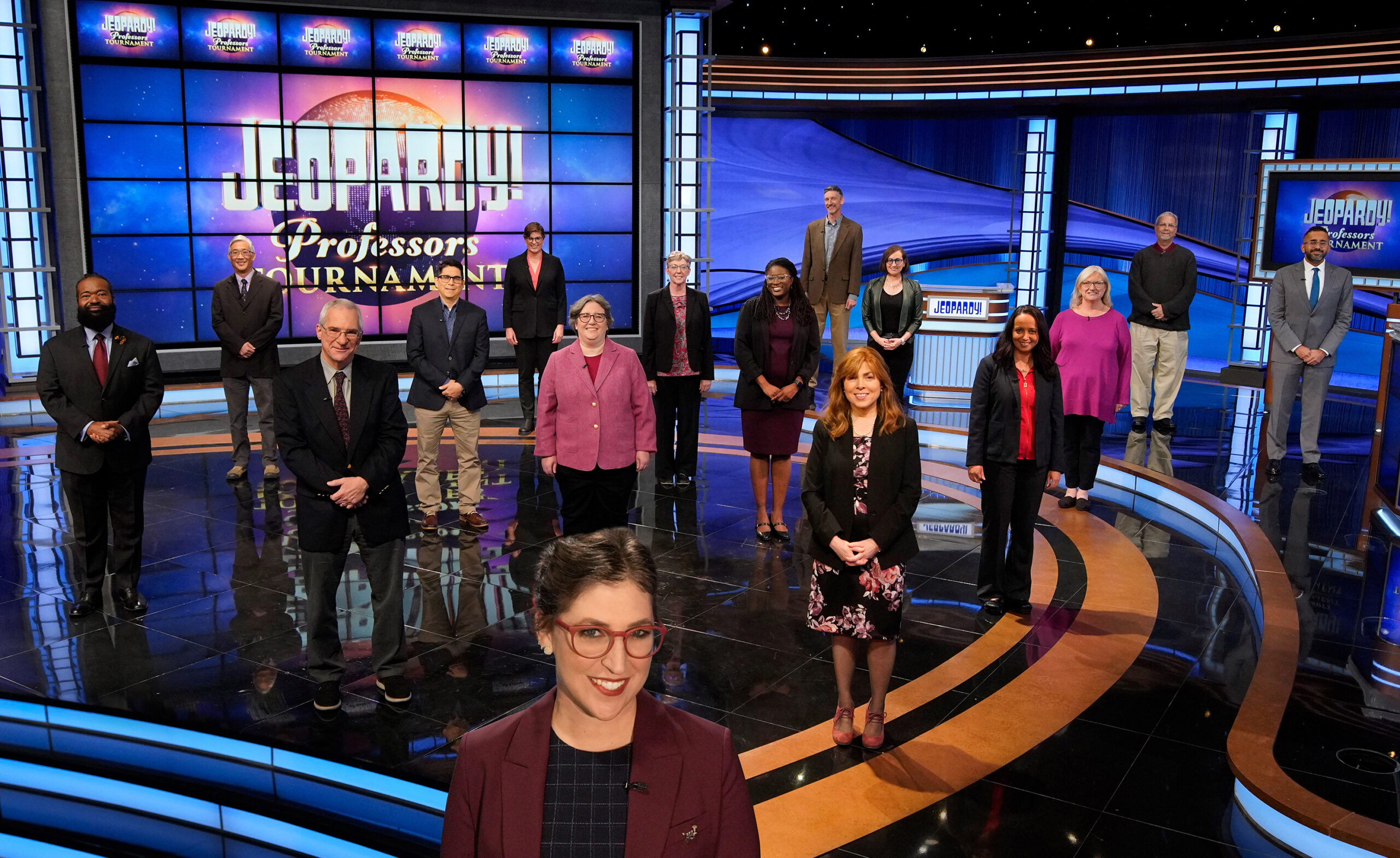Jeopardy! host Mayim Bialik standing on set in front of the contestants of the Professors Tournament.